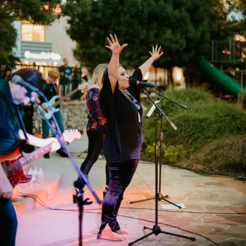 The image shows a lively outdoor musical performance. In the foreground, a woman is passionately singing into a microphone, raising both arms high with open hands. She is dressed in a black outfit and is standing on a stone path. In the background, a band is playing with several musicians holding instruments like guitars. One musician in the foreground is playing an electric guitar. The setting appears to be an outdoor area with greenery and mature trees, creating a casual and inviting ambiance. Lights cast a warm glow on the performers, enhancing the atmosphere of the event. A small crowd is visible in the background, gathered around the performance space.