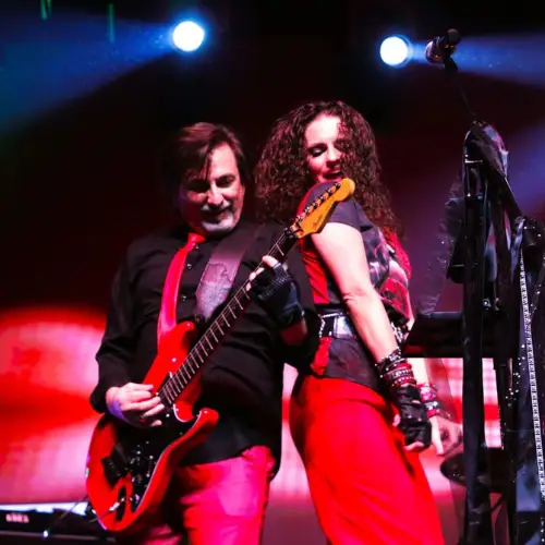 The image shows two musicians performing on stage. The man on the left is playing a bright red electric guitar and is dressed in a black shirt and shiny red pants. He appears focused on his performance. A woman stands to his right, leaning back against him playfully, smiling, and holding a keyboard. She wears a graphic T-shirt and vibrant red pants, accessorized with numerous bracelets and a belt. The background is dark with stage lights illuminating them, creating a dramatic and vibrant atmosphere. A microphone stands in front of them on the stage.