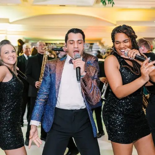 A lively scene with three people in sparkly outfits singing and dancing at an event. Bright lighting and a cheering crowd create an energetic atmosphere.