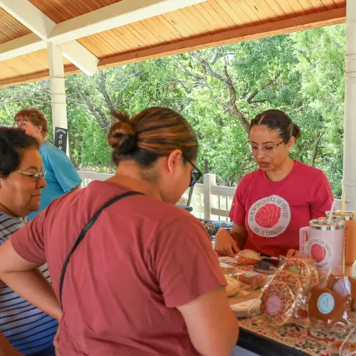 A vendor wearing a shirt that says "In a world full of donuts, be a concha" hands baked goods to a customer at an outdoor market stall.