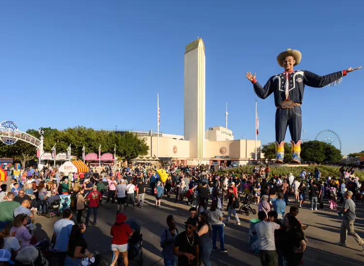 Crowds at the Texas State Fair with Big Tex statue and fair attractions in the background.
