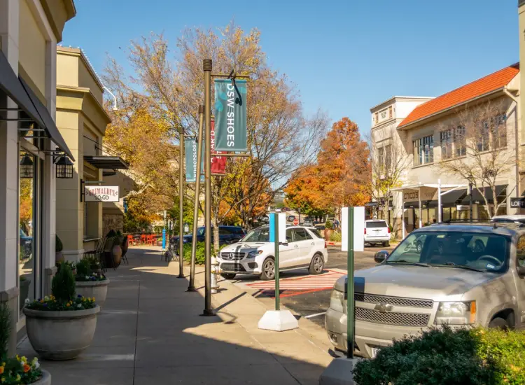 Watters Creek shopping village street with stores, cars, and autumn trees in Allen, Texas.