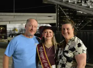 Teen wearing sash and cowboy hat poses with two adults at a community event at night