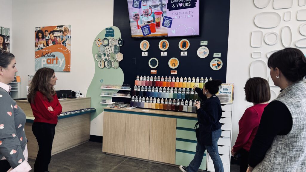 Guests gathered inside Color Me Mine in Allen, Texas, listening as a staff member explains glaze colors and painting techniques in front of a wall display filled with paint bottles and sample ceramics.