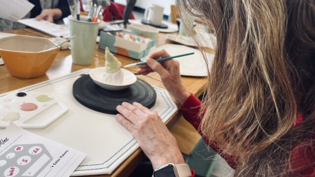 Guest painting a ceramic gnome figurine during a pottery painting session at Color Me Mine in Allen, Texas, with glaze colors, brushes, and tools arranged on a wooden table.