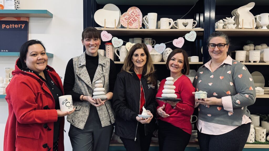 Group of women posing inside Color Me Mine in Allen, Texas, holding hand-painted ceramic pieces during a pottery painting experience, with shelves of unfinished ceramics and seasonal décor in the background.