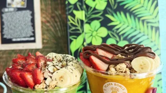 Two smoothie bowls topped with strawberries, bananas, granola, and chocolate drizzle held in front of a tropical-themed wall at Playa Bowls in Allen, Texas