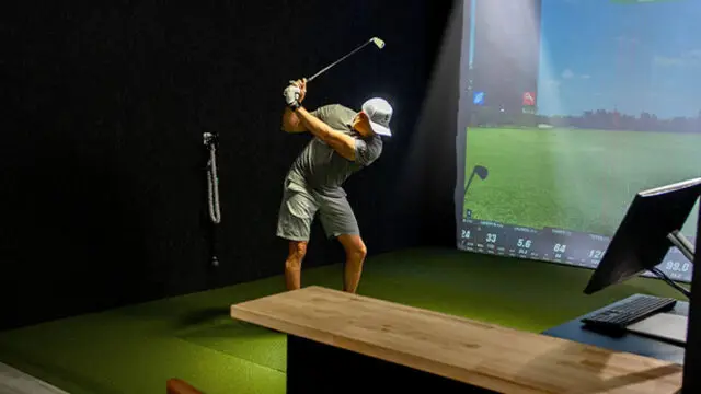 Man swinging a golf club in an indoor simulator bay with projected course screen and swing analysis data