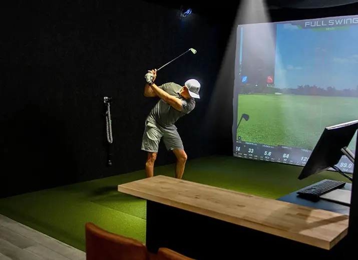 Man swinging a golf club in an indoor simulator bay with projected course screen and swing analysis data