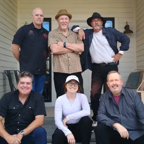 Members of The Last Exit band posing together on a porch, with three standing and three seated on the steps.
