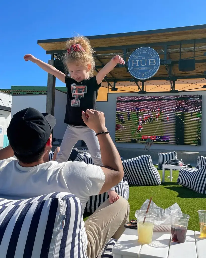 Child playing on outdoor seating area while watching a game on a large screen at The HUB in Allen, Texas.