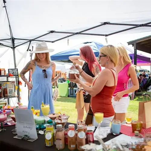 Women shopping from a vendor booth with drinks, candles, and handmade goods at an outdoor market.