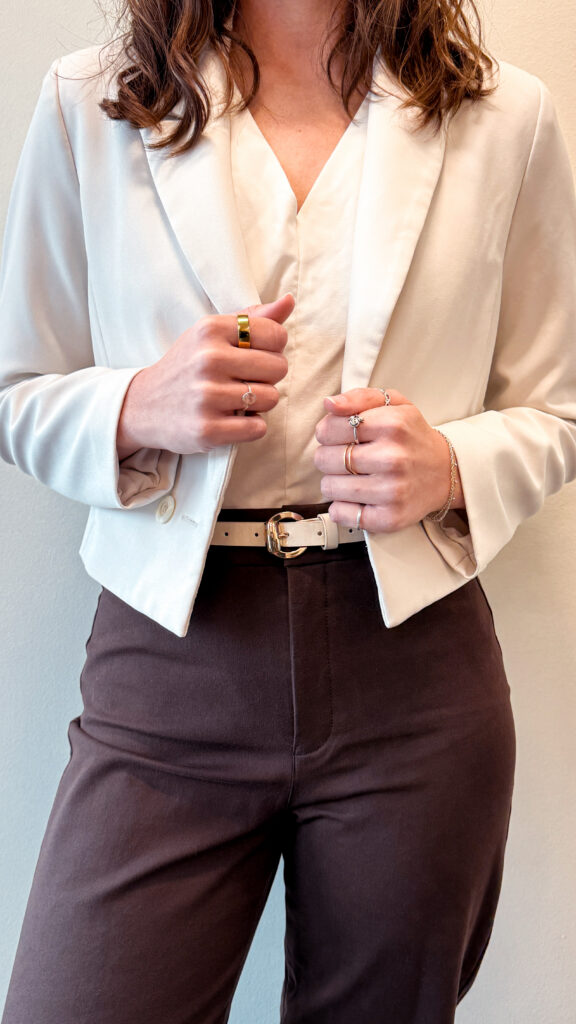 Person wearing a cream blazer, neutral blouse, and brown trousers styled with layered jewelry and a light belt.