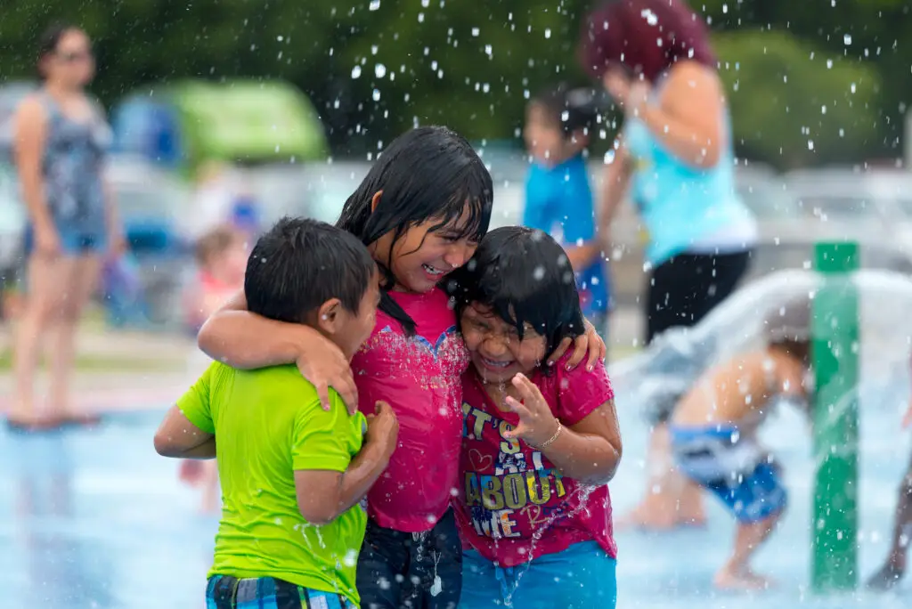 Three children hugging and laughing while playing in water sprays at an outdoor splash pad in Allen, Texas.