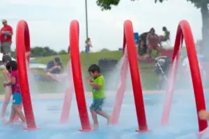 Children running through water sprays at a colorful outdoor splash pad with red arches in a public park.