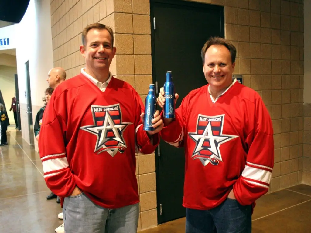 Two men wearing red Allen Americans hockey jerseys hold Bud Light bottles inside an arena concourse.