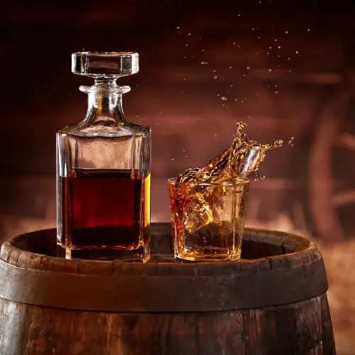 Glass decanter and whiskey tumbler with a splash of amber bourbon displayed on a wooden barrel during a spirits tasting experience.