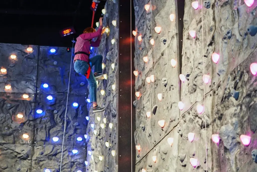 Person climbing a colorful indoor rock wall attraction at PINSTACK in Allen, Texas.