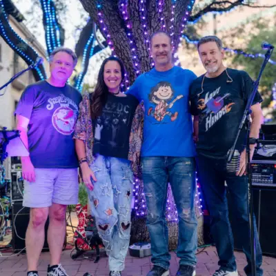 Members of the band Love Handle posing together outdoors in front of a tree wrapped with colorful lights and live music equipment.