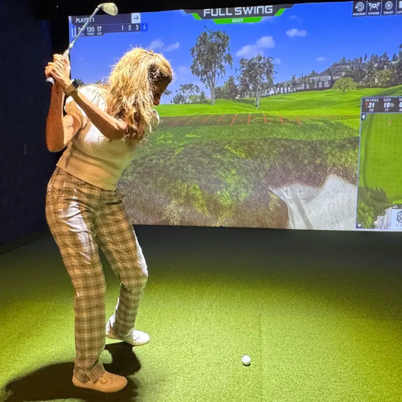 Woman swinging a golf club inside The Back Nine indoor golf simulator with a virtual golf course projected on a large screen.