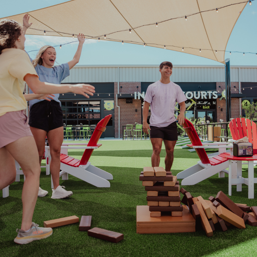 Group of friends playing a giant block stacking game on the outdoor lawn patio at Chicken N Pickle in Allen, Texas.