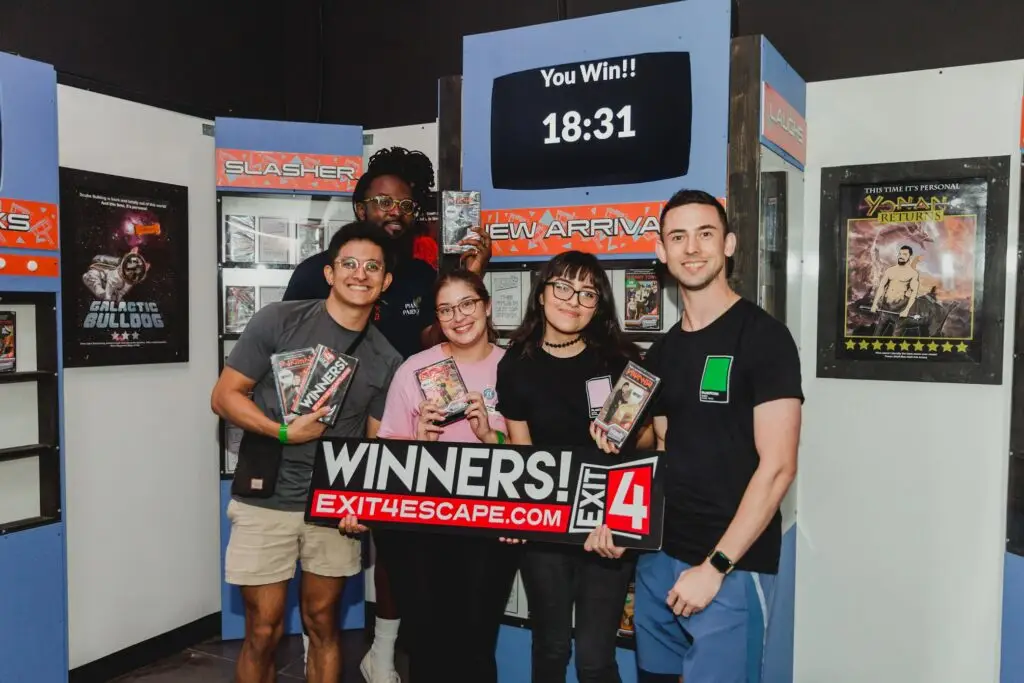 A group of five guests celebrates escaping a room at EXIT4 Escape Rooms in Allen, Texas, holding a winners sign and game-themed prizes beneath a screen displaying their completion time.