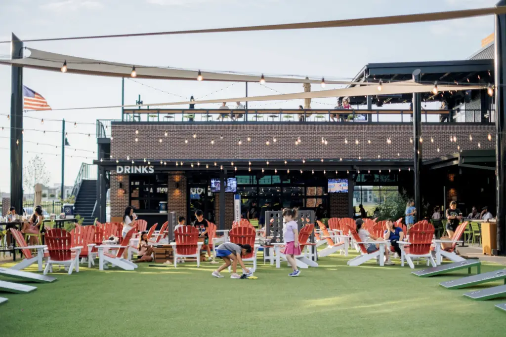 Families and groups enjoy the outdoor lawn, games, and patio seating at Chicken N Pickle in Allen, Texas, with string lights and the two-story venue creating a lively summer atmosphere.