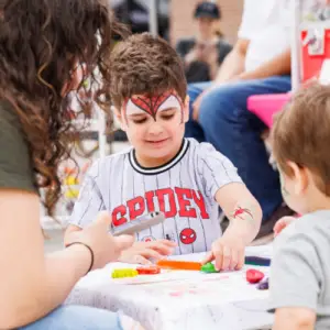Child wearing a Spider-Man shirt and face paint coloring at an activity table during a family-friendly outdoor event while other children and adults participate nearby.