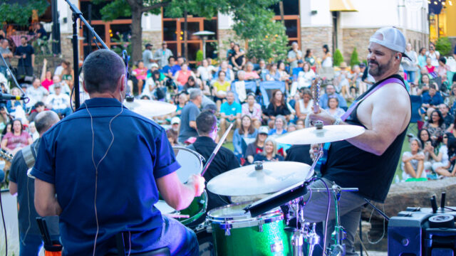 View from the stage of a live band performing at Concerts by the Creek while a large crowd gathers on the lawn at Watters Creek Village.