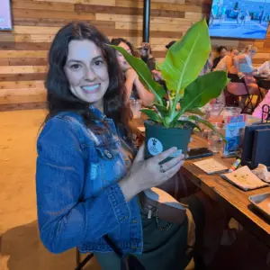 A smiling woman holds a potted tropical houseplant she won during Plant Bingo at Chicken N Pickle in Allen, Texas, with bingo cards, food, and other guests enjoying the event in the background.