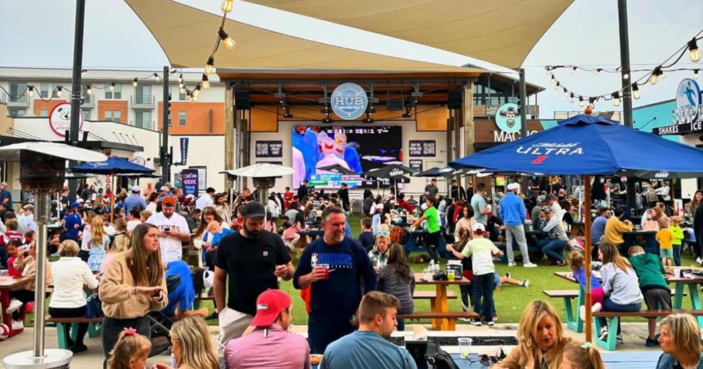 Crowds gather at The HUB in Allen, Texas, watching entertainment on a large outdoor screen while enjoying food, drinks, and family-friendly activities.