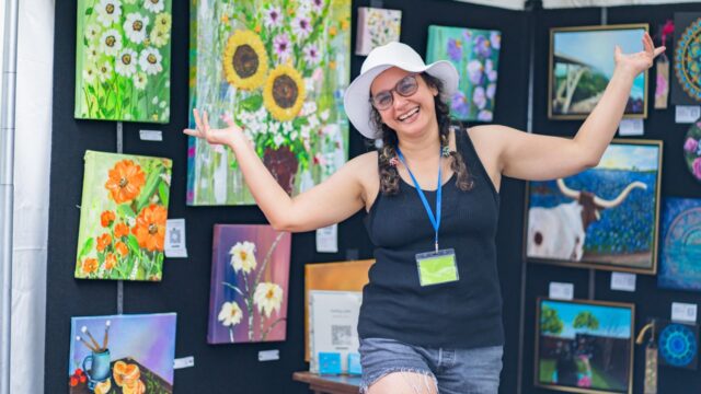 Artist standing in front of a colorful booth display of paintings and artwork at the Allen Arts Festival at Watters Creek Village.