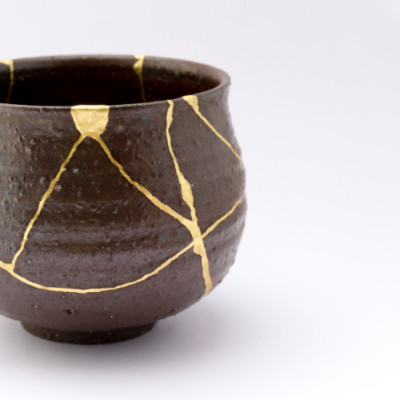 Close-up of a handcrafted ceramic cup repaired using the Japanese art of kintsugi, with gold-filled cracks creating a decorative pattern across the dark pottery against a light background.