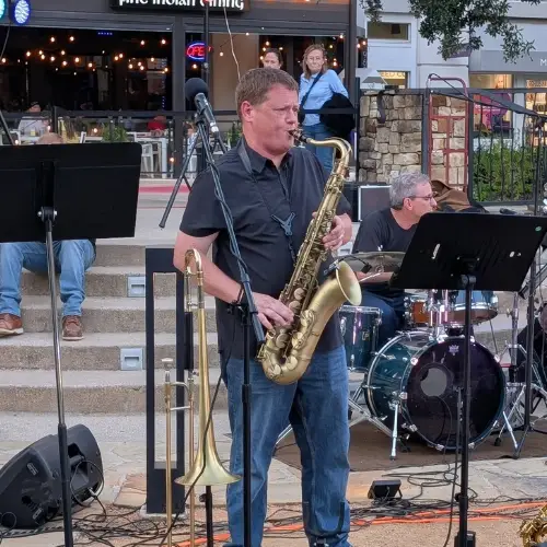 A saxophone player performs with Last Call featuring The Top Shelf Brass during Concerts by the Creek at Watters Creek Village in Allen, Texas, as the band entertains guests outdoors.