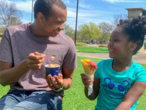 An adult and child enjoy ice cream together at The Village at Allen in Allen, Texas, smiling as they share a treat outdoors on a sunny day.