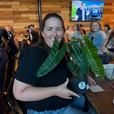 Guest smiles while holding a potted houseplant during a plant bingo event at Chicken N Pickle in Allen, Texas, with other participants seated in the background.