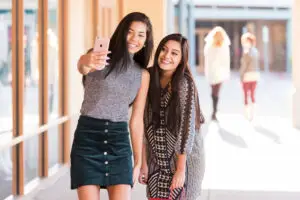 Two friends take a selfie while shopping at Allen Premium Outlets in Allen, Texas, with storefronts and other shoppers visible in the background.