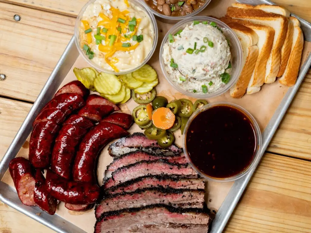 A barbecue platter from Vaqueros Texas Bar-B-Q in Allen, Texas, featuring sliced brisket, smoked sausage, macaroni and cheese, potato salad, beans, pickles, jalapeños, toast, and barbecue sauce.
