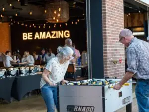 Couple plays foosball together at Chicken N Pickle in Allen, Texas, with other guests and a glowing “Be Amazing” sign in the background.