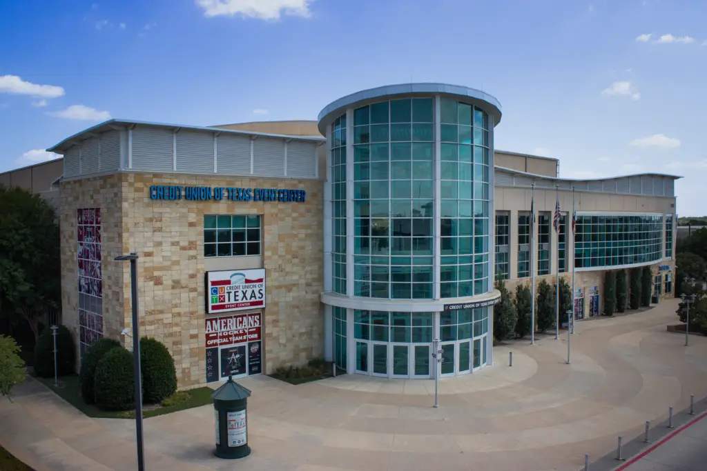 Exterior of Credit Union of Texas Event Center in Allen, featuring its curved glass entrance, stone façade, plaza and venue signage under a blue sky.