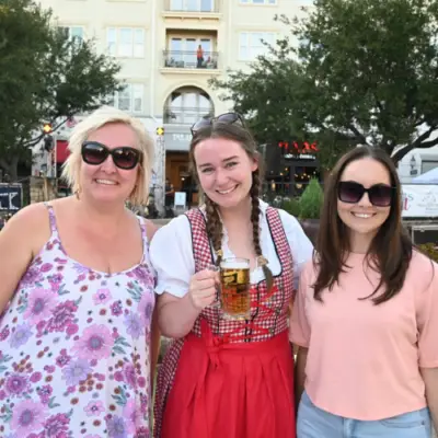 Three women smile at the Crafts & Drafts Fine Arts Festival at Watters Creek Village in Allen, Texas, with one guest holding a beer during the outdoor event.