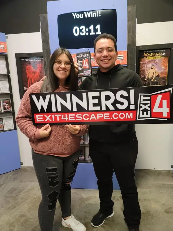 Smiling couple holds a “Winners!” sign after successfully completing an escape room at Exit 4 Escape Rooms in Allen, Texas.