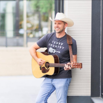 Bryan Mayberry smiles while playing acoustic guitar and wearing a white cowboy hat and Texas Country T-shirt.