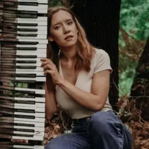 Carly Carroll poses beside a vertically positioned keyboard in a wooded outdoor setting.