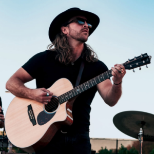Clayton Stewart performs on acoustic guitar while wearing a black cowboy hat and sunglasses.