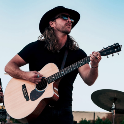 Clayton Stewart performs on acoustic guitar while wearing a black cowboy hat and sunglasses.