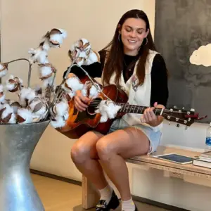 Courtney Istre smiles while seated and playing an acoustic guitar beside a display of cotton stems.