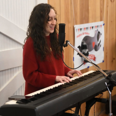 Emmeline sings into a microphone while playing keyboard during an indoor performance.