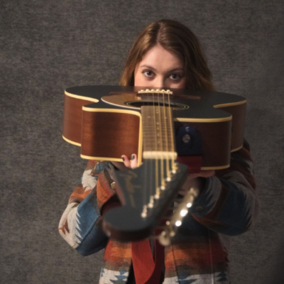 Jacy Penland holds an acoustic guitar toward the camera in a playful musician portrait.