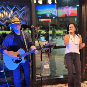 Rima sings into a microphone while Tommy plays acoustic guitar during a live indoor performance.